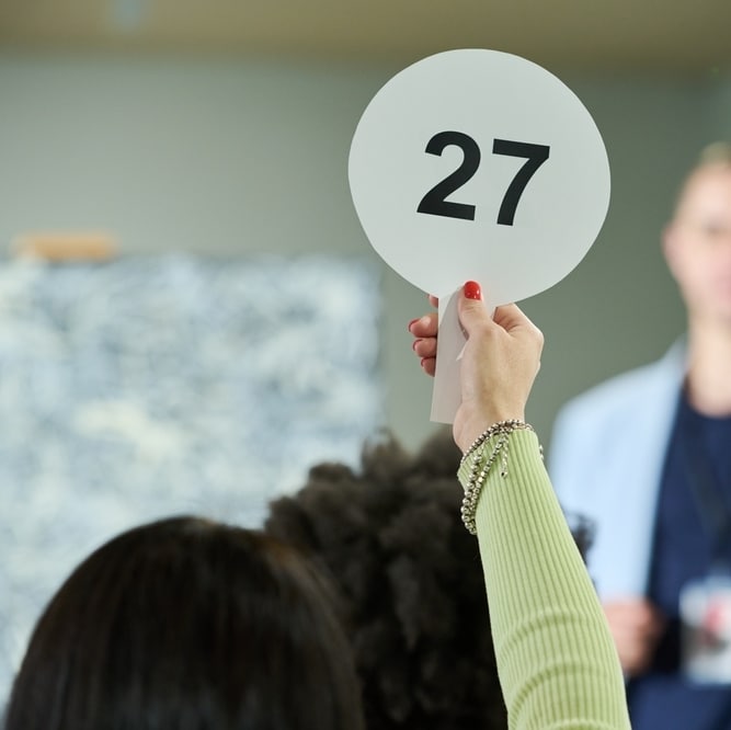Enchères Publiques : Main levant le Panneau Numéro 27 Main levant une plaque d'enchères numéro 27 lors d'une vente publique, avec manche vert et bracelets.